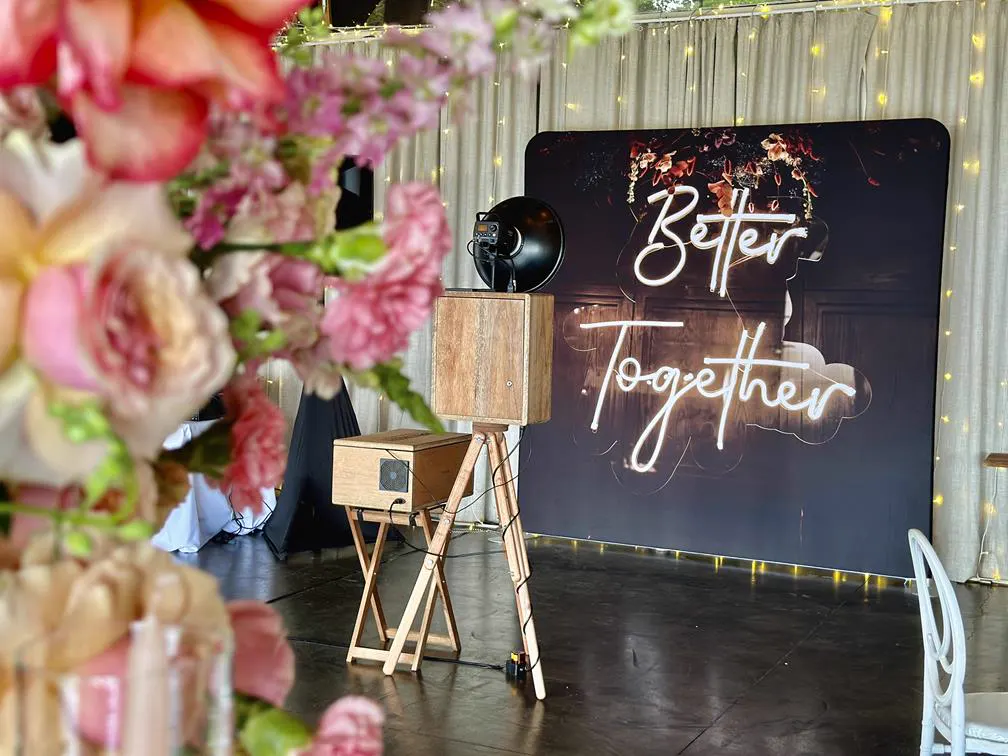 The Retrobooth facing a Better Together neon backdrop framed by pink and coral wedding florals